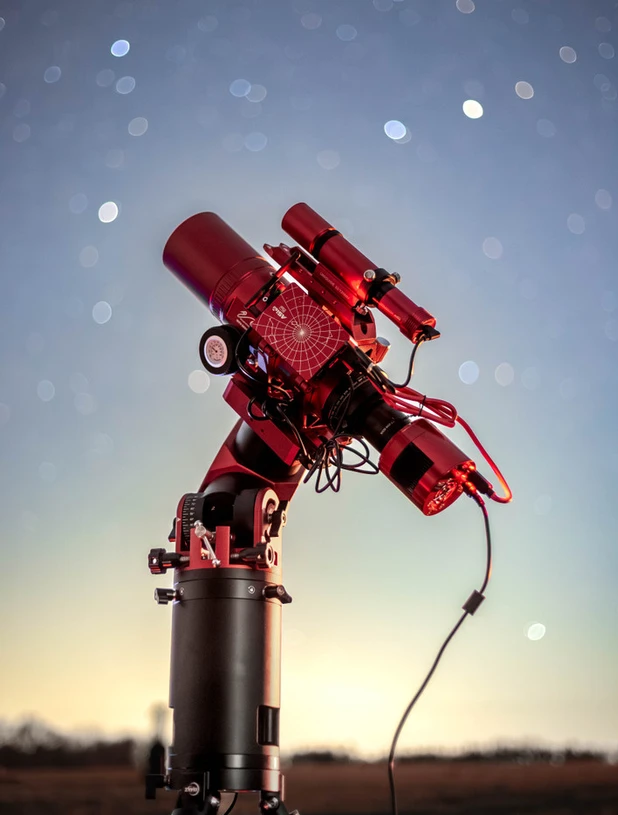 Red-lit telescope and camera equipment on an equatorial mount beneath a starry sky.